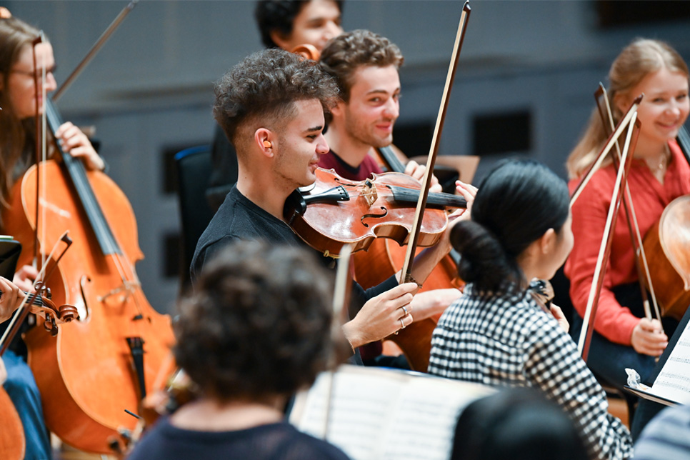 A group of string musicians, sitting together in an orchestra rehearsal, smiling and chatting to each other.